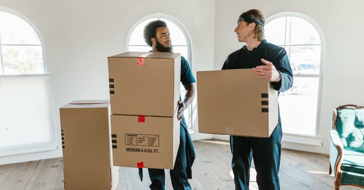 Dos personas transportando grandes cajas de cartón en una habitación luminosa con ventanas arqueadas.