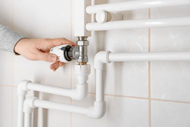 Hand turning a valve on white bathroom pipes against a tiled wall