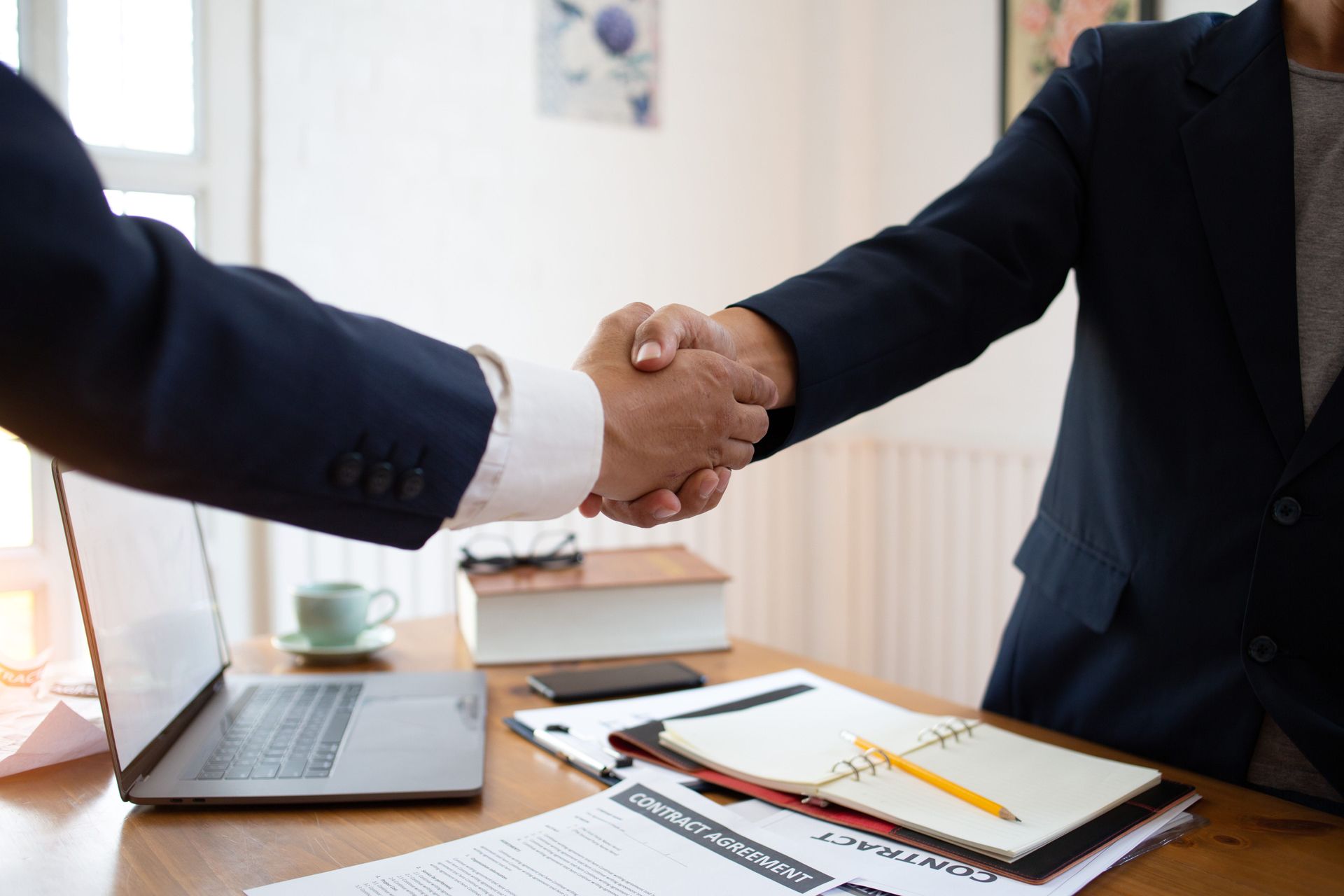 Two businesspeople shaking hands across a desk with a laptop and documents