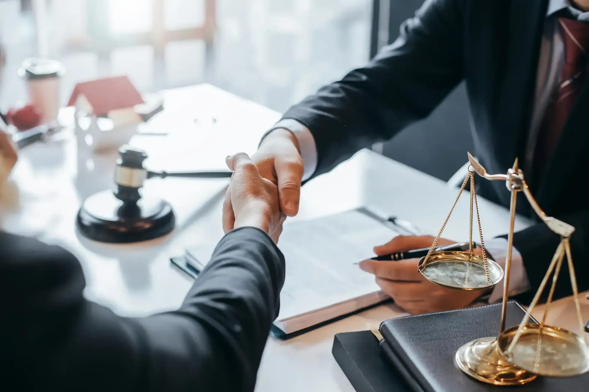 Two people in suits shaking hands at a desk with scales of justice and a gavel.