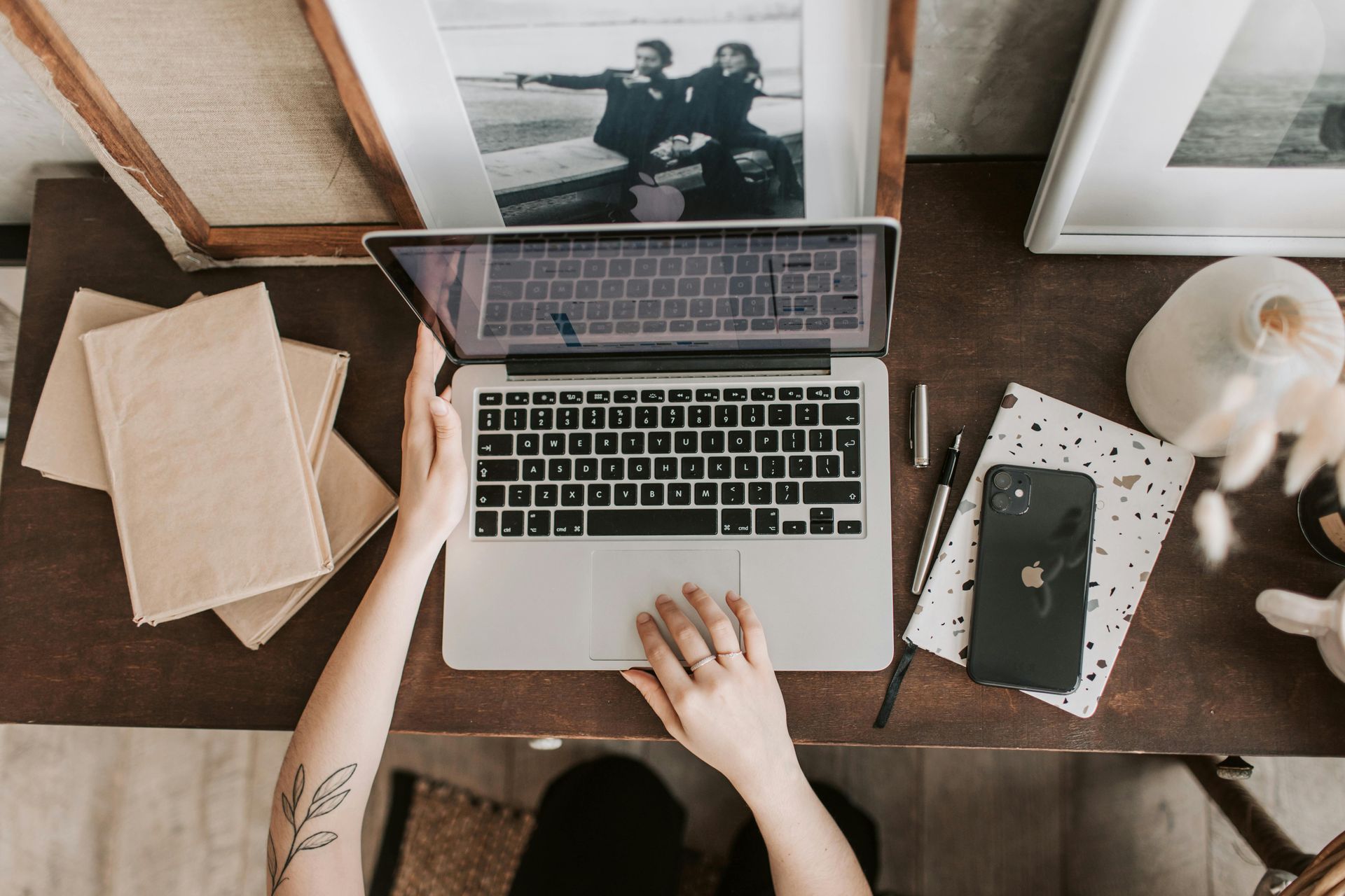 Person using laptop at a desk with books, a phone, and framed photos.