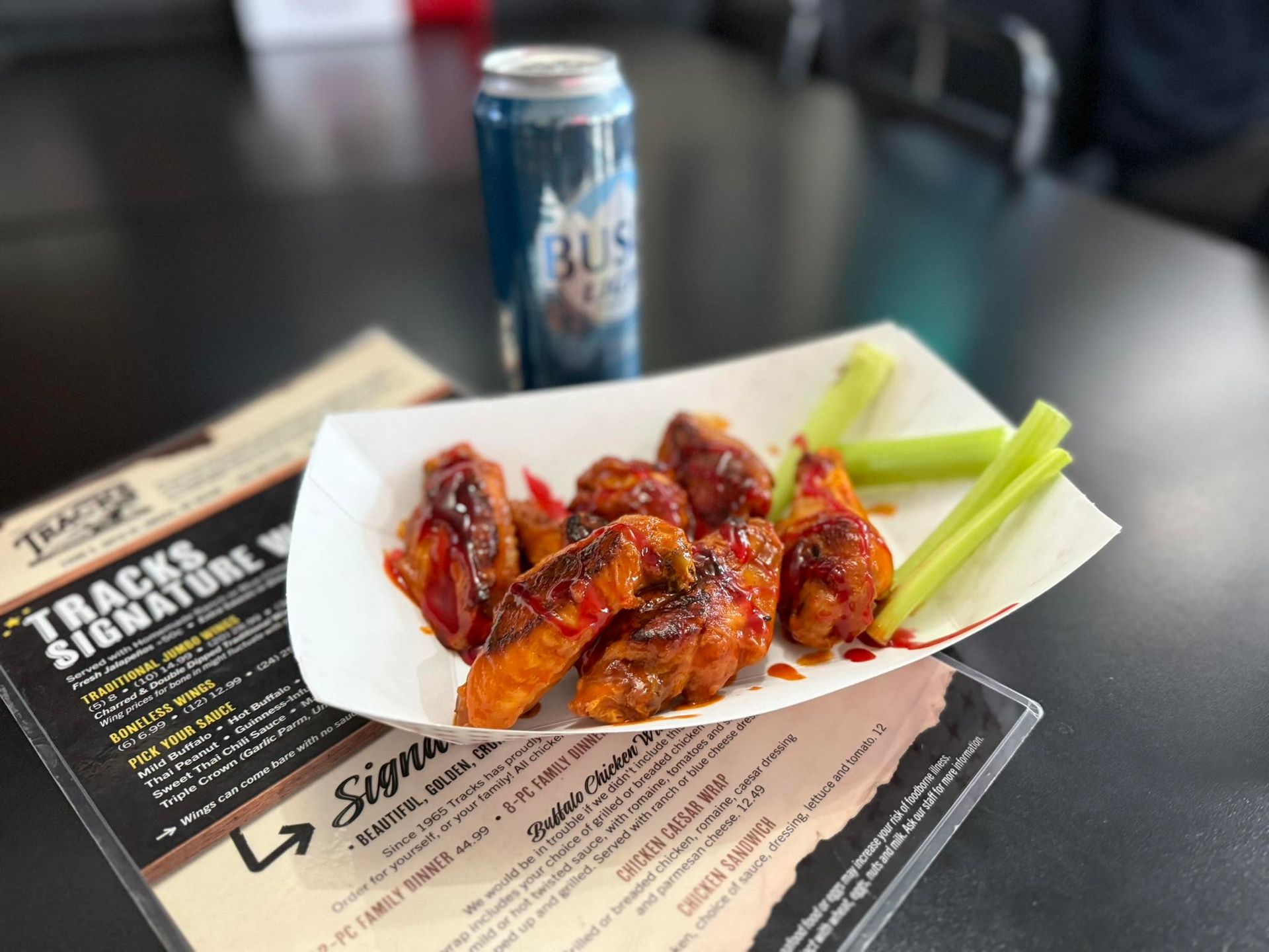 Tray of glazed chicken wings with celery on a table beside a can of Busch beer