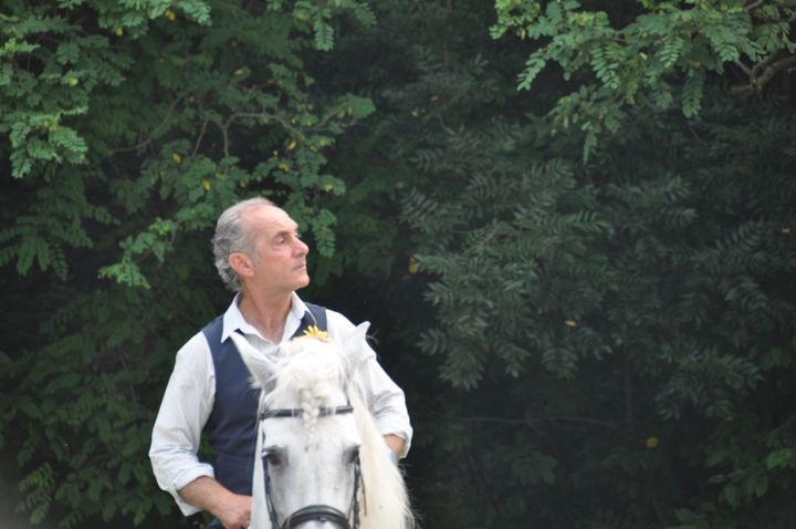 Uomo con camicia bianca e gilet scuro a cavallo di un cavallo bianco, con uno sfondo verde e frondoso