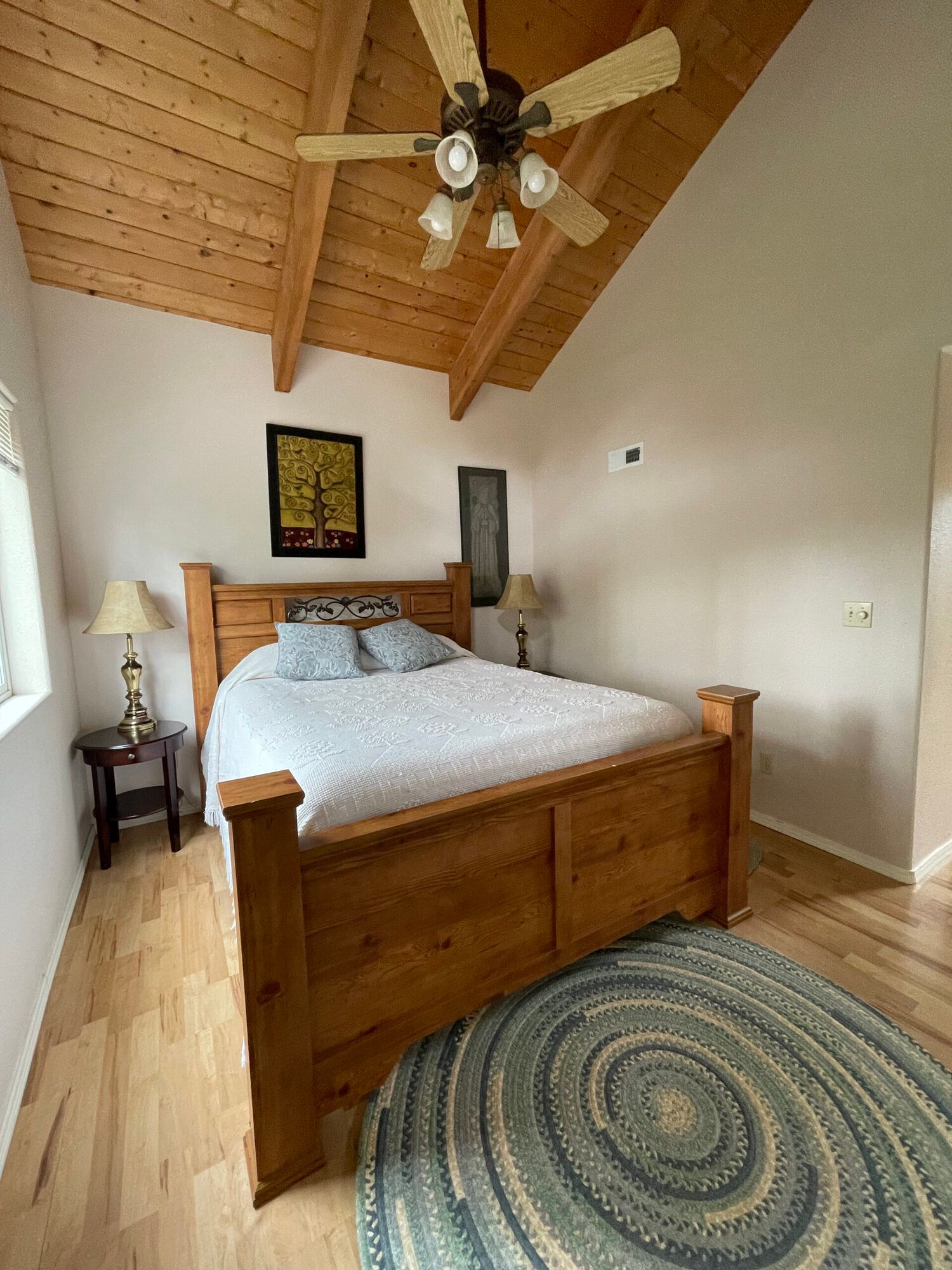 A bedroom with a wooden bed frame, light wood floors, a vaulted wood ceiling, ceiling fan, and a round blue rug.