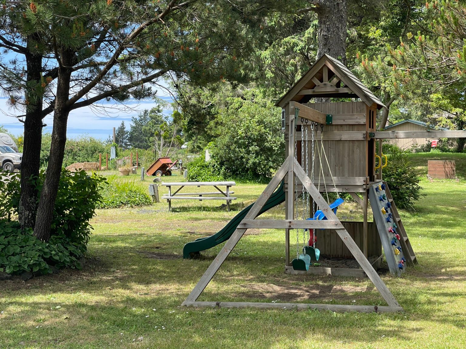 A wooden playset with a green slide and a climbing wall sits in a grassy park area near a picnic table and trees.