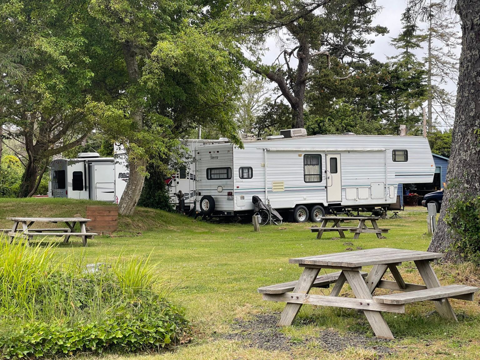 Two large white travel trailers parked in a grassy campground with wooden picnic tables in the foreground.
