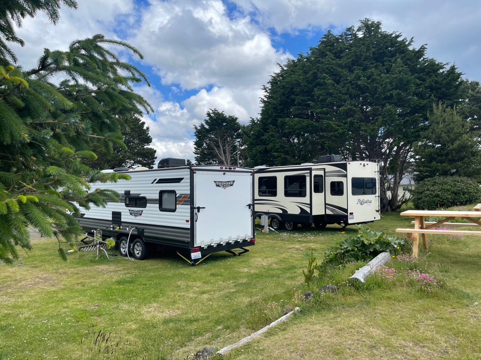 Two travel trailers parked on a grassy campground under a cloudy sky, with a wooden picnic table nearby.