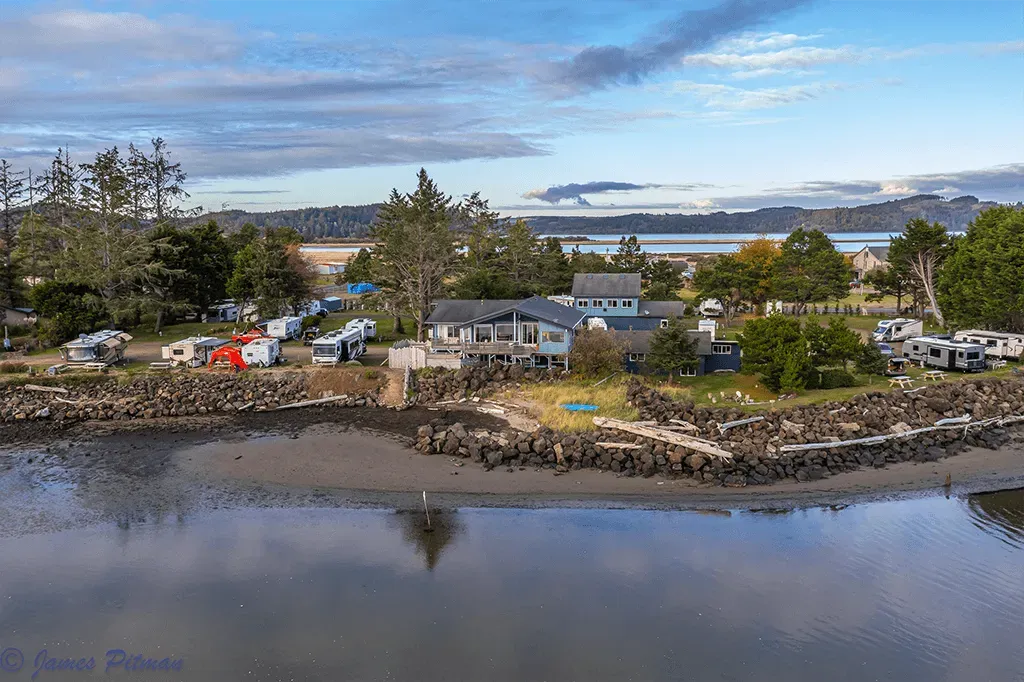 A waterfront campground with RVs, a central house, and many trees bordering a river under a cloudy blue sky.