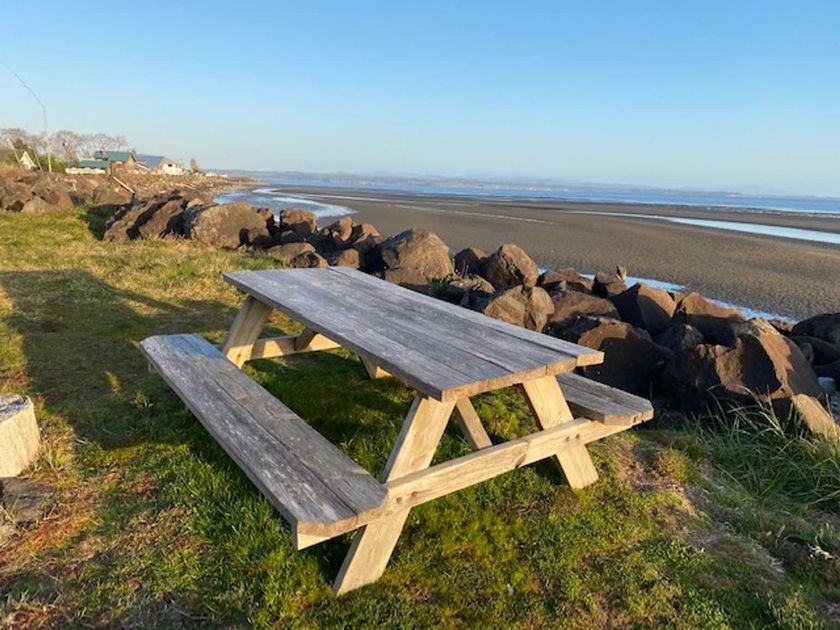 A weathered wooden picnic table sits on a grassy bank overlooking a wide, sandy beach and the ocean under a clear sky.