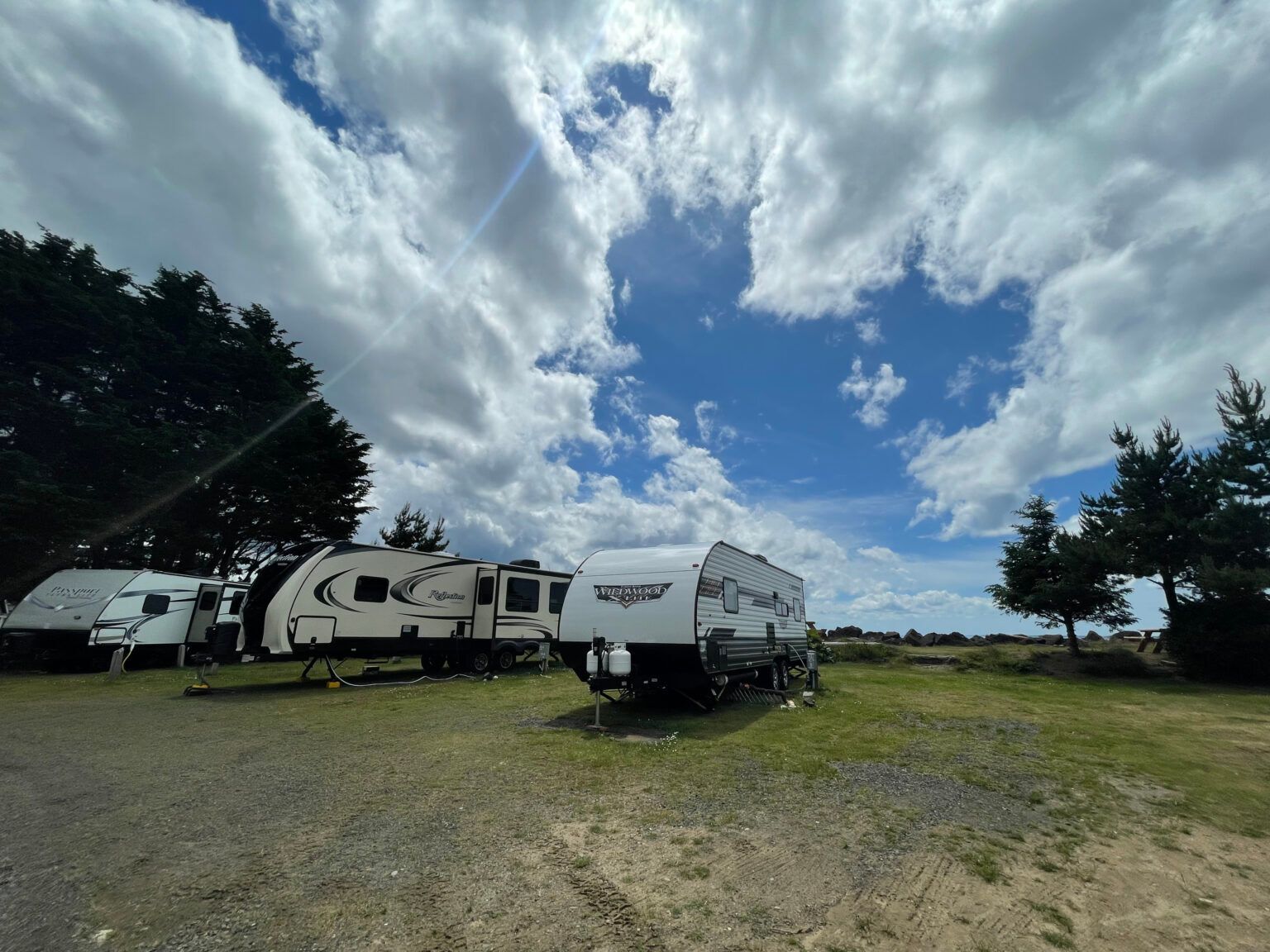 Two travel trailers parked on a gravel lot under a bright, partly cloudy blue sky.