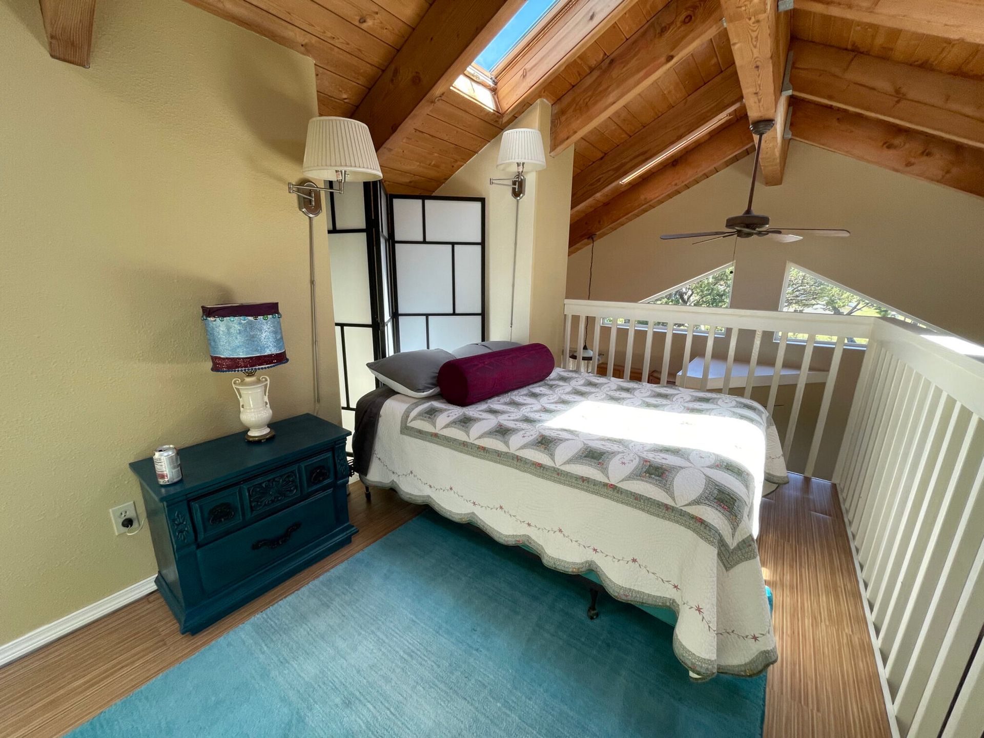 A bedroom in a loft with a bed, a blue nightstand with a lamp, a blue rug, and wooden beams on the slanted ceiling.