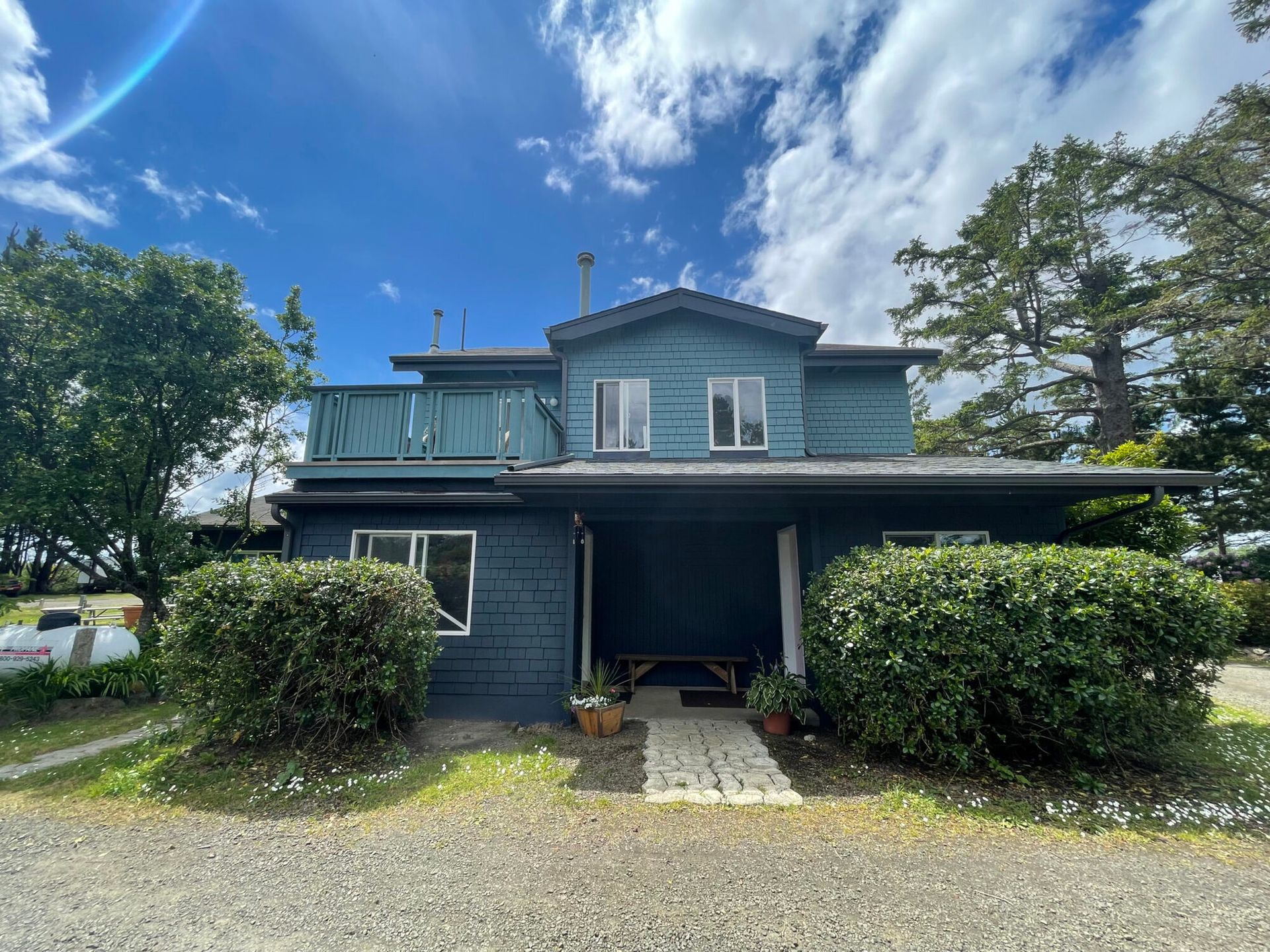 A two-story blue house with a balcony and dark green bushes in the front, set against a bright, cloudy sky.
