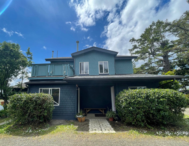 A two-story blue house with a balcony, a front porch, and large green bushes under a bright, partly cloudy blue sky.