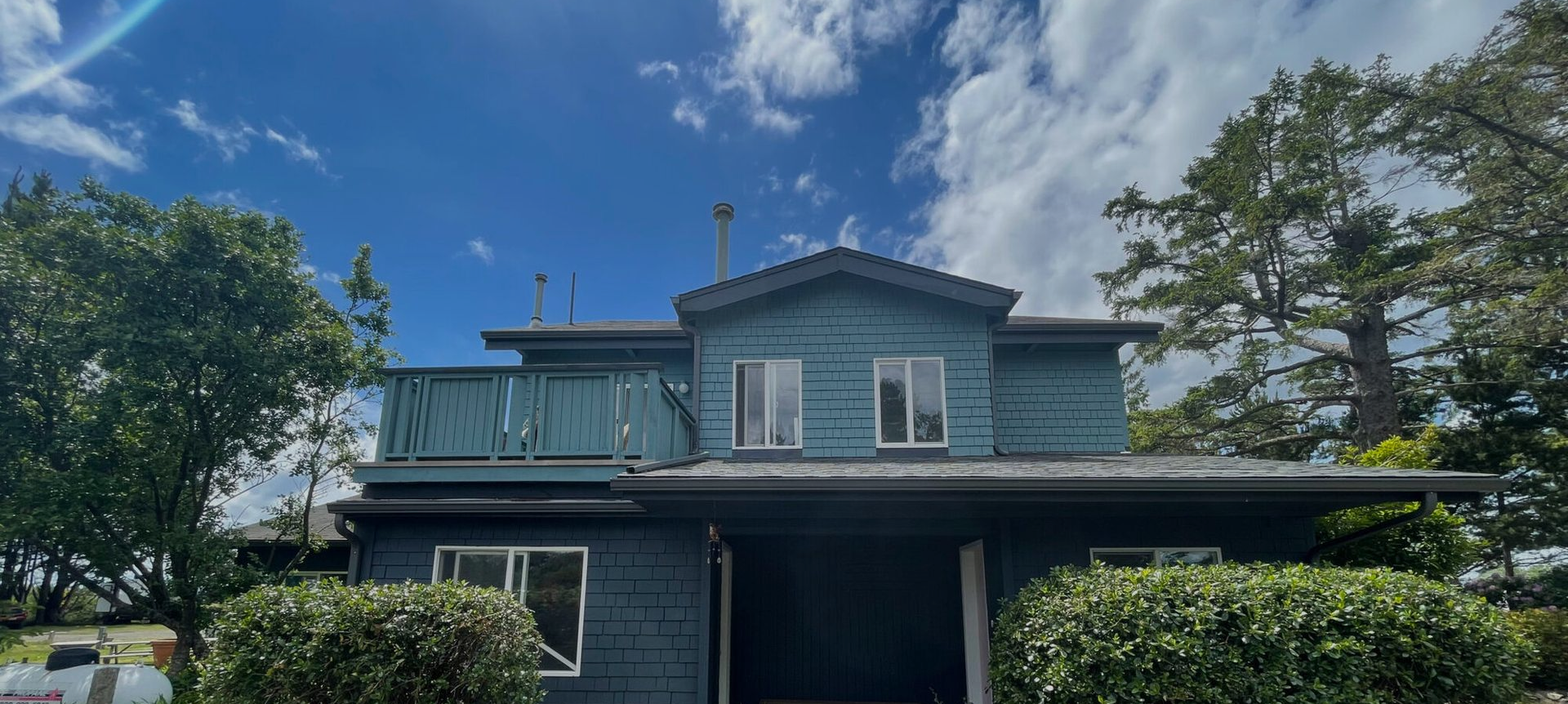 A two-story blue house with a balcony and dark wood siding, surrounded by green trees under a bright blue sky.