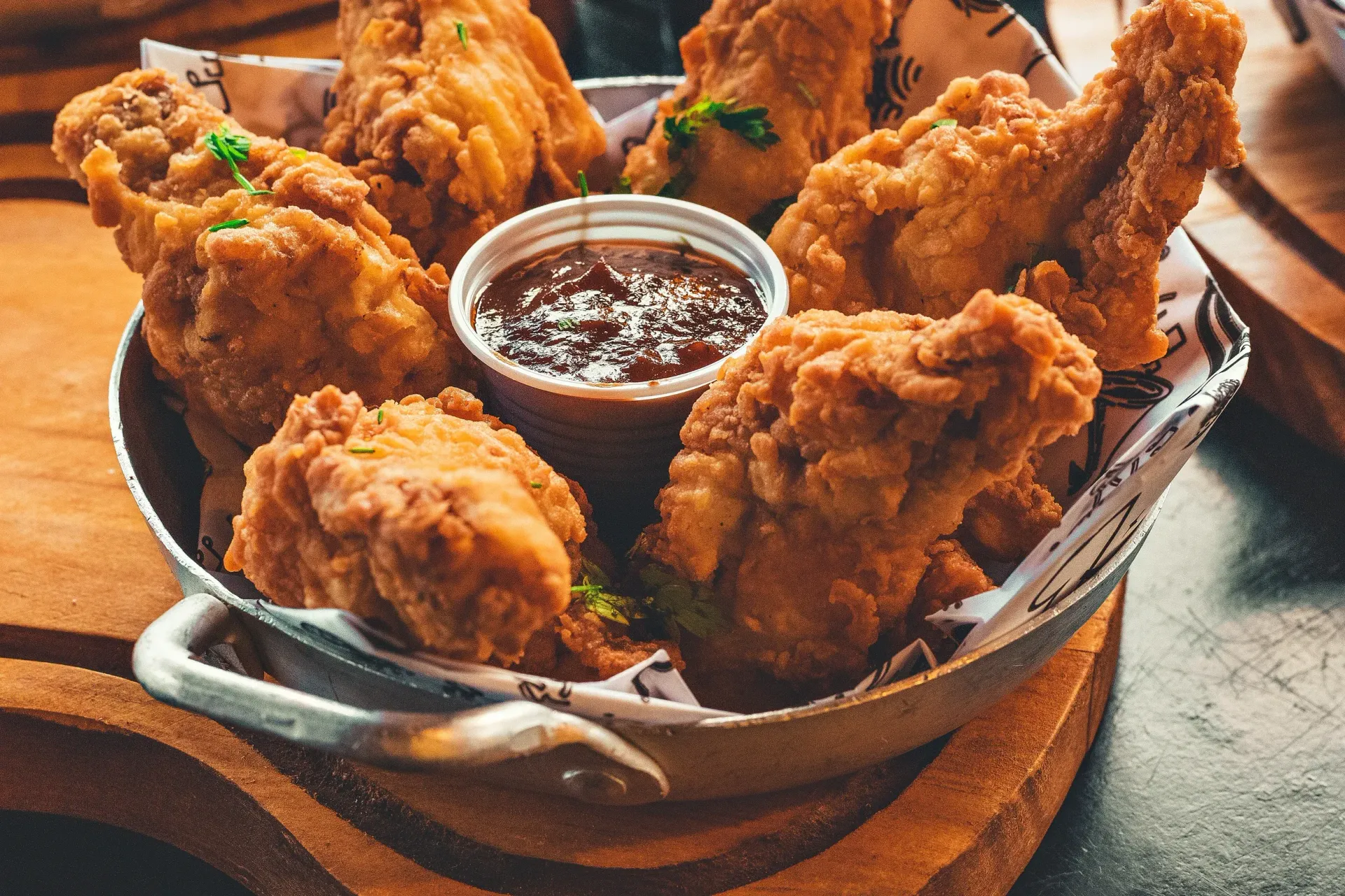 A metal basket filled with golden-brown fried chicken pieces and a small container of dark dipping sauce on a wooden board.