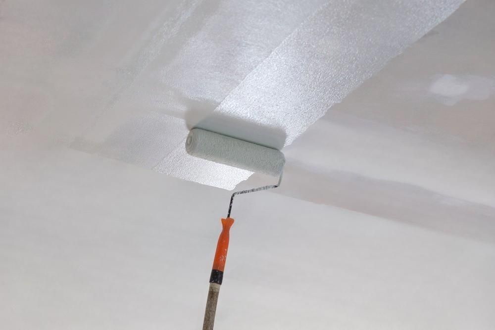 A paint roller applies white paint to a white ceiling.