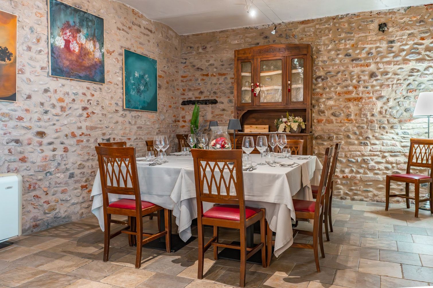 Accogliente sala da pranzo in stile rustico con tavolo apparecchiato, sedie in legno, pareti in pietra e opere d'arte colorate.