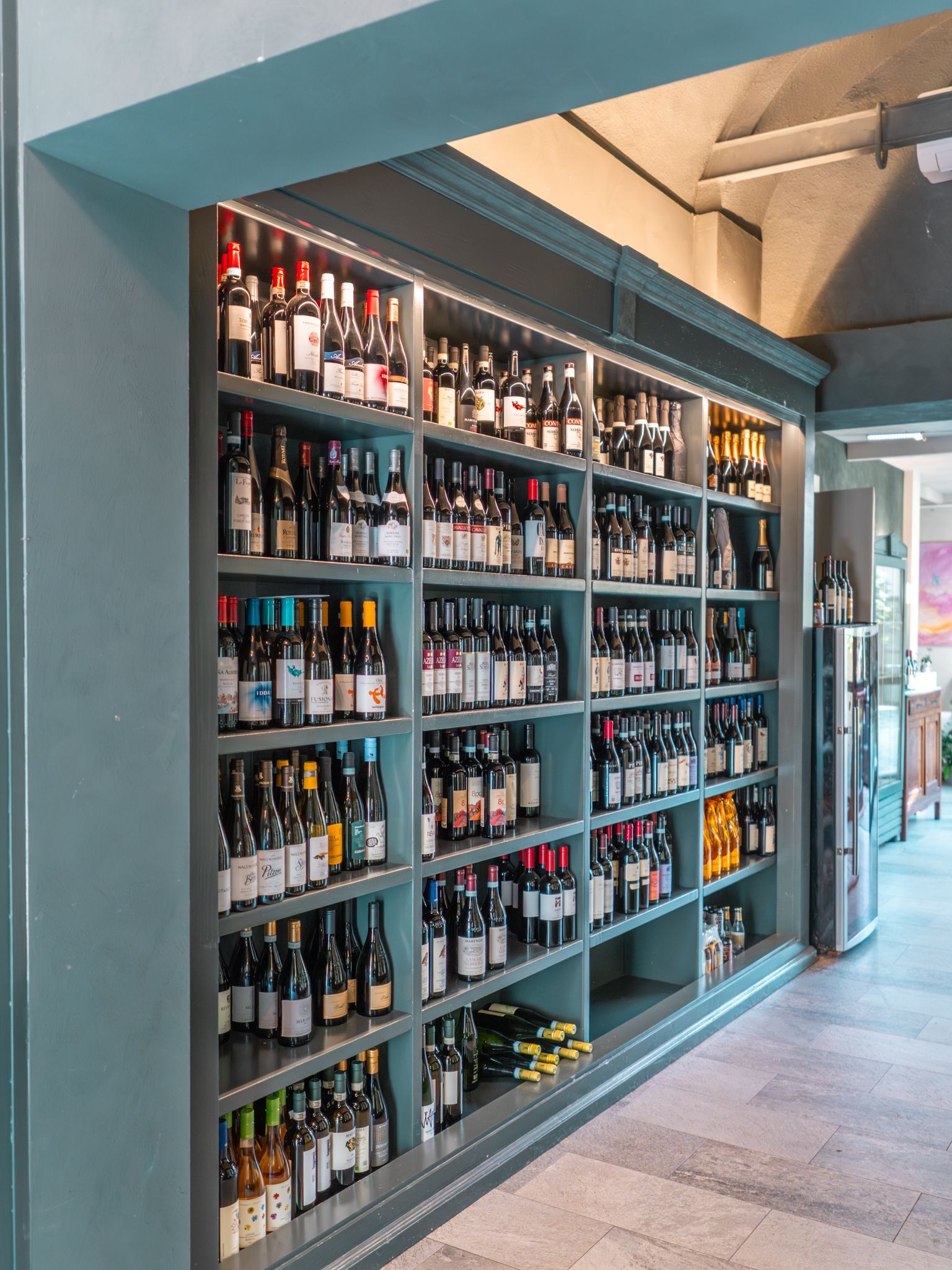 Bottiglie di vino esposte su scaffali nel corridoio di un negozio o di un ristorante con pareti color verde acqua.