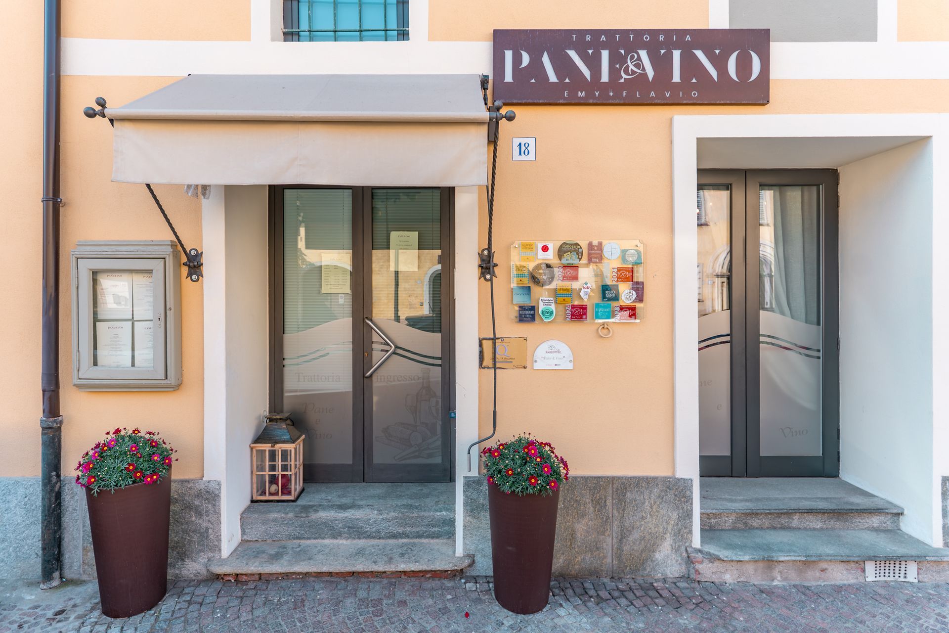 Ingresso di PANATTO con porte a vetri, vasi di fiori e bacheca su una facciata color pesca.