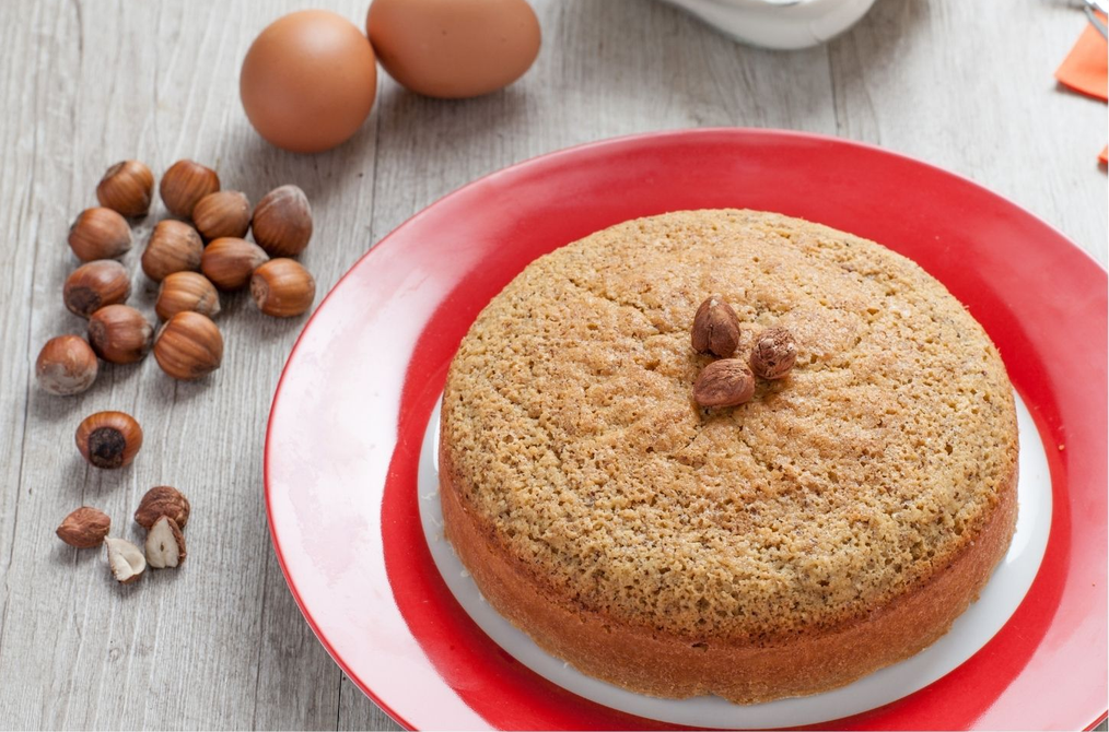 Torta rotonda su un piatto rosso con nocciole e due uova su un tavolo di legno chiaro.