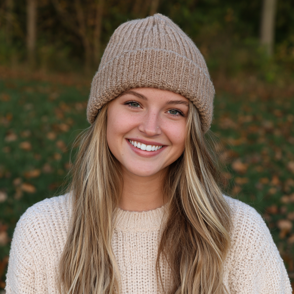 Smiling person in a beige knit hat and sweater outdoors with blurred autumn foliage behind.