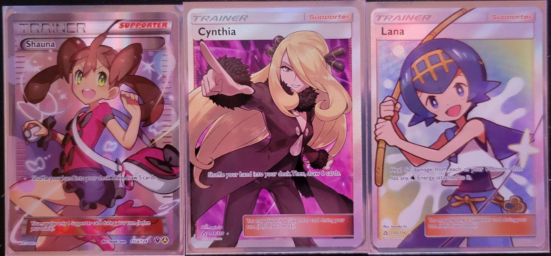 three Pokémon full Art Trainer cards sitting next to each other in a row