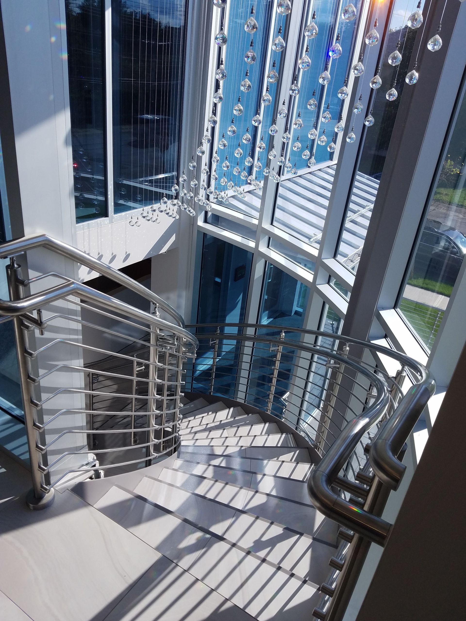 Curved metal stairway beside tall glass windows, with sunlight casting striped shadows indoors.