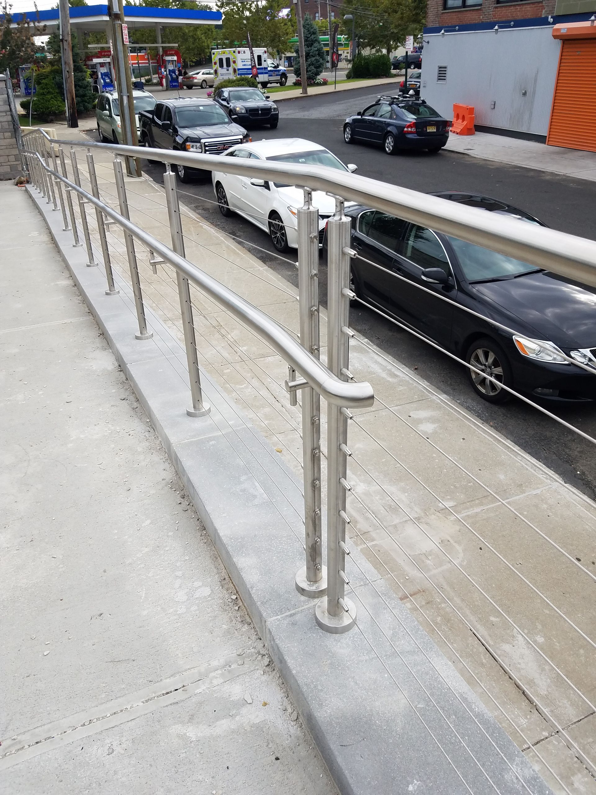 Sidewalk ramp with metal handrails beside a street, with parked cars and buildings in the background.