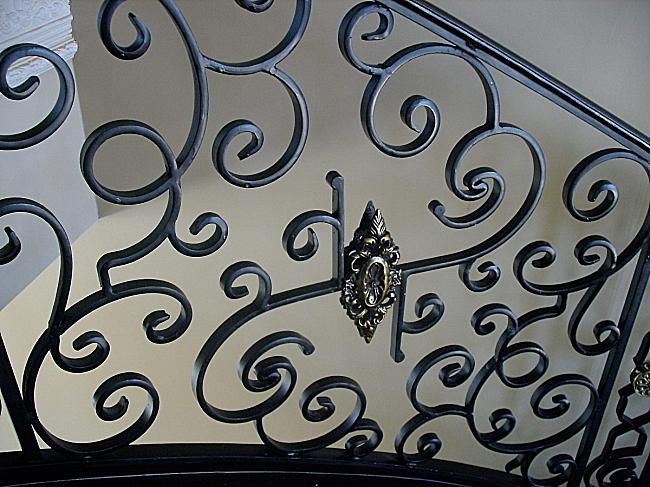 Black wrought-iron decorative gate with ornate swirling scrollwork and a central latch against a light wall.