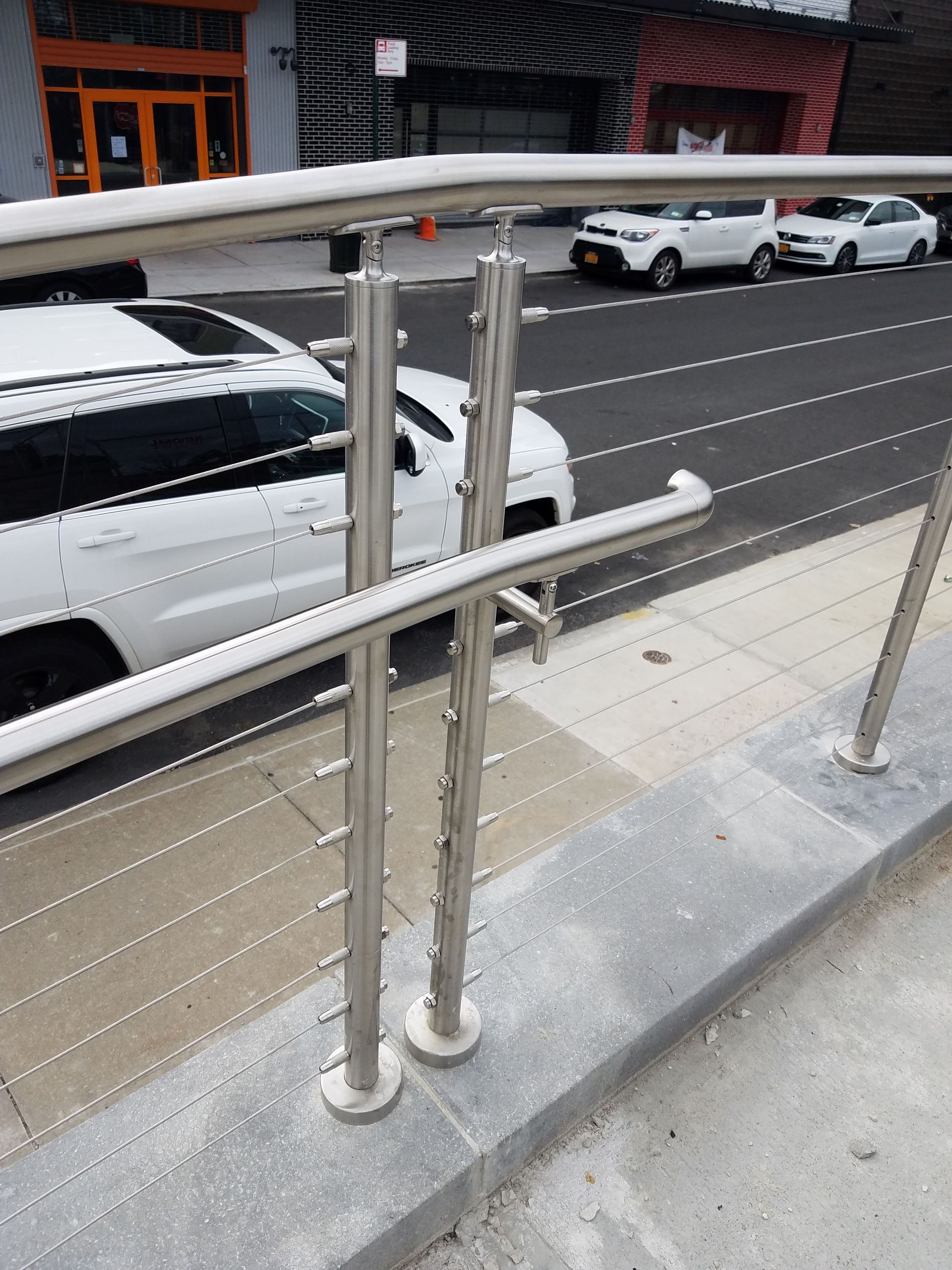 Metal railing and sidewalk beside a street with parked cars and a white van near buildings