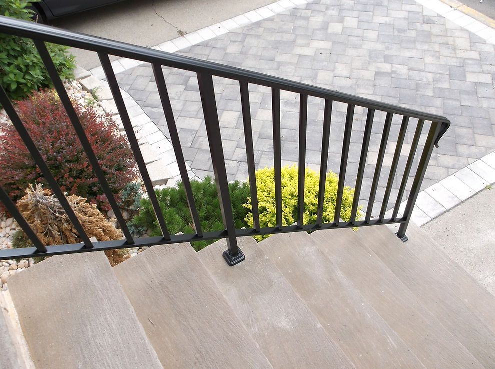 Black metal stair railing beside tan steps and a paved walkway with landscaping below