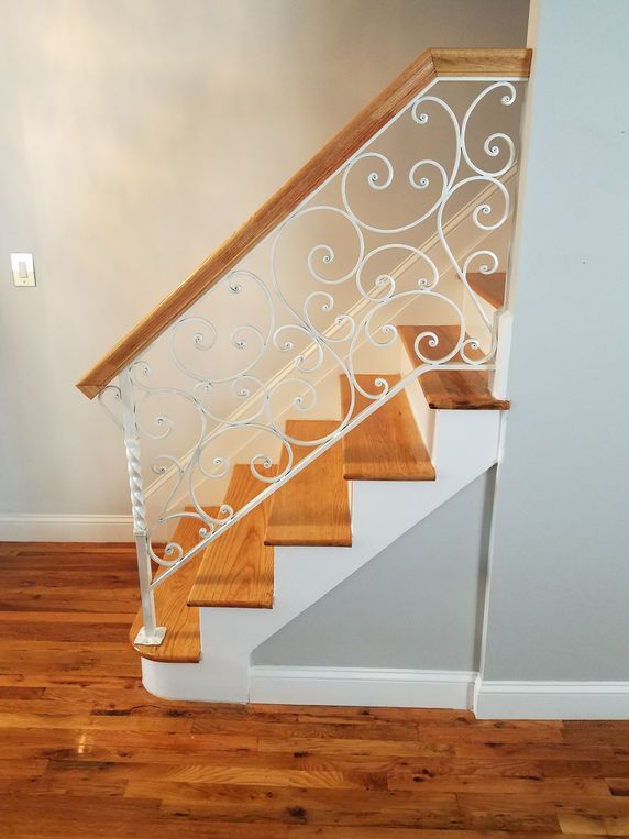 Wooden staircase with white decorative wrought-iron railing beside a light gray wall and hardwood floor