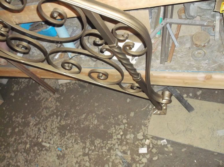 Ornate metal stair railing with scrollwork beside a dusty concrete floor and wall framing materials