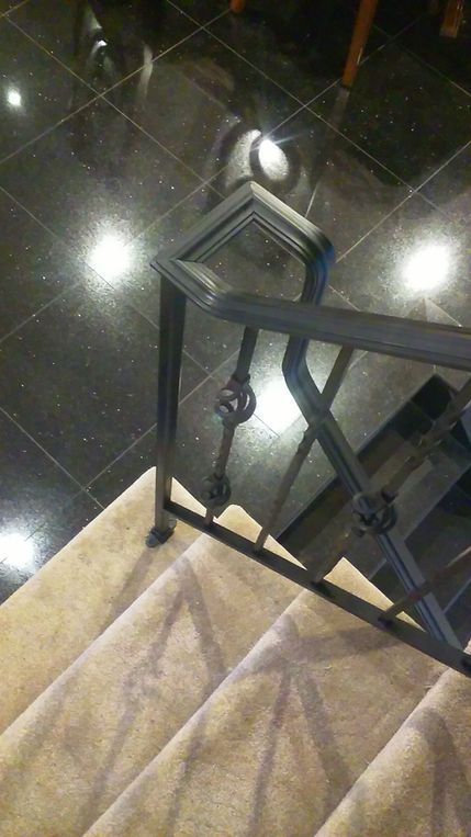 Staircase landing with black metal railing, beige tiled steps, and shiny dark floor reflecting ceiling lights
