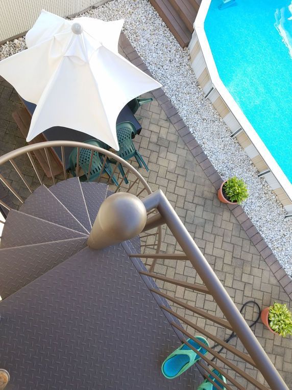 Overhead view of a poolside patio with a white umbrella, brown chairs, potted plants, and a blue pool edge