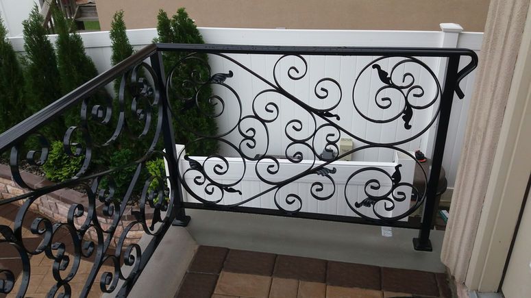 Black wrought-iron balcony railing with ornate scrollwork beside potted evergreens on a tiled patio