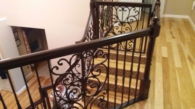 Ornate black wrought-iron staircase railing beside a light wood floor and carpeted stairs.