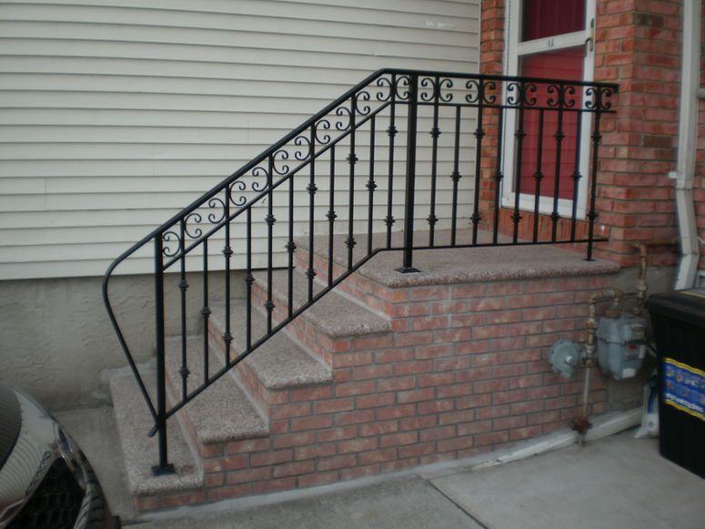 Front steps with black wrought-iron railing beside a brick house and red door