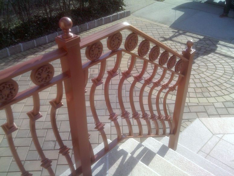 Brown metal railing with wavy decorative bars along an outdoor paved walkway in sunlight