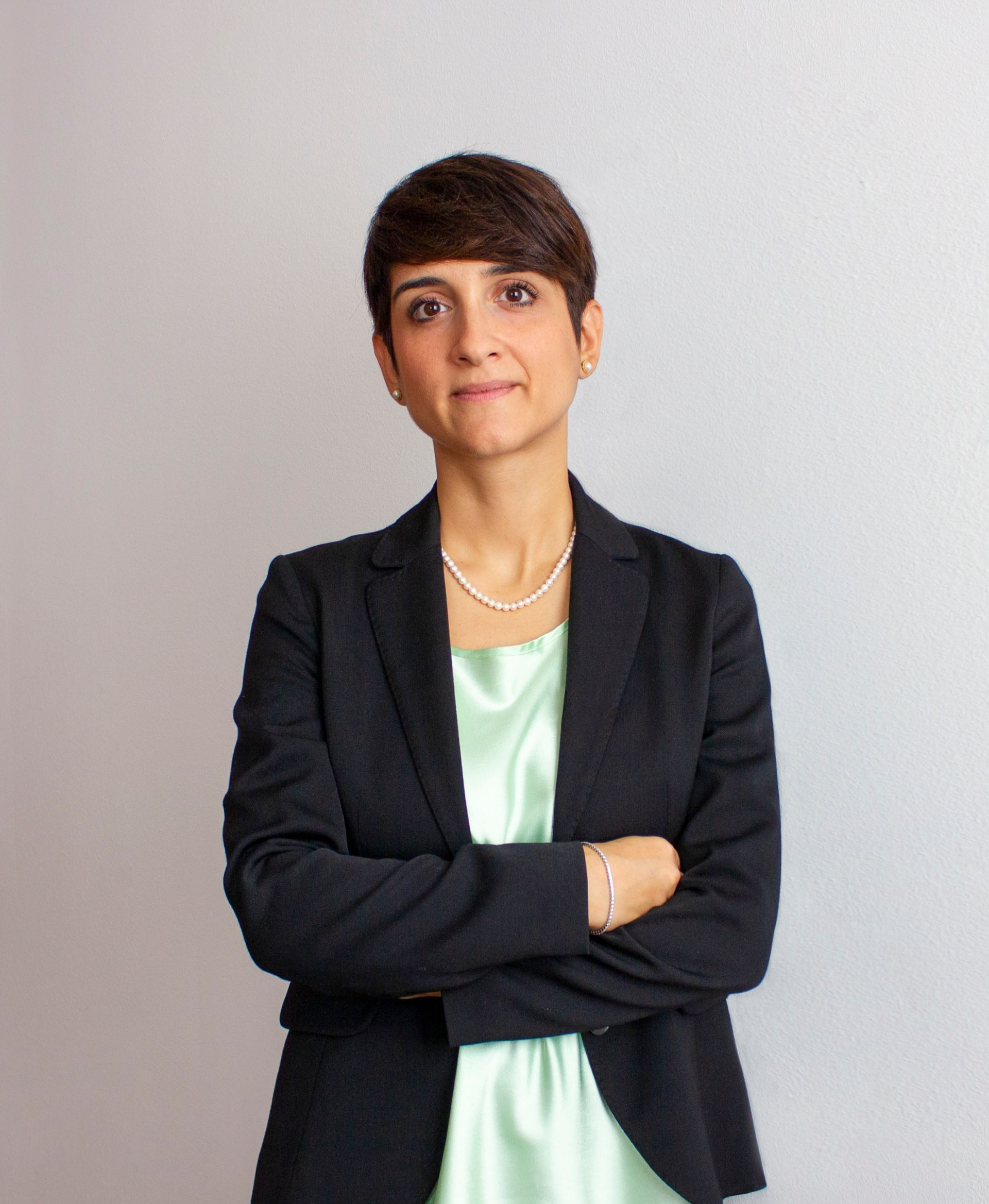 Persona in piedi con le braccia incrociate, che indossa un blazer nero sopra una maglietta color menta su sfondo grigio.