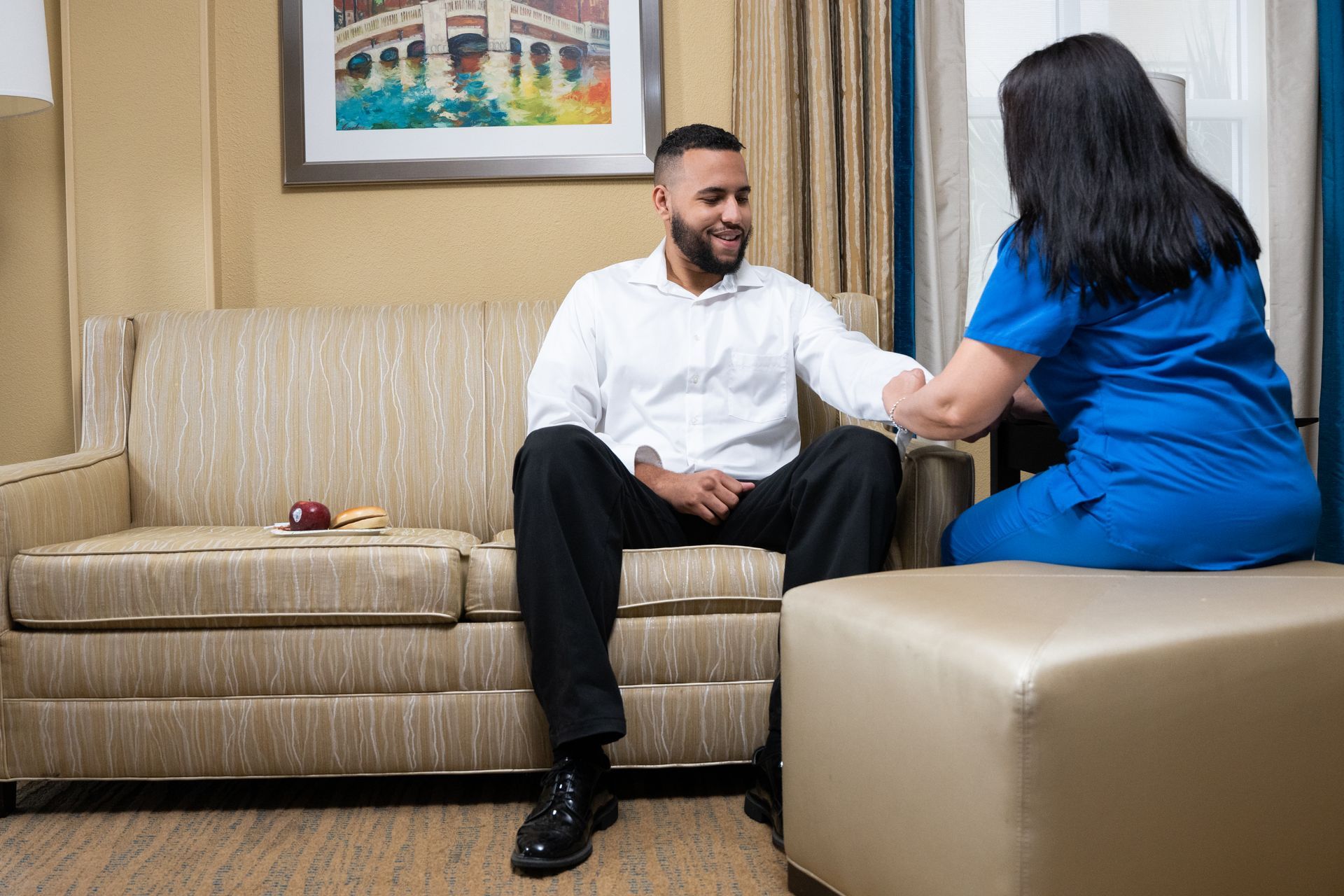 Two people talking on a beige couch in a hotel room, one in a white shirt and one in a blue top.