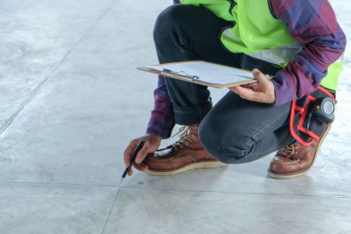Worker kneeling on concrete floor, marking a line with a pencil while holding a clipboard and wearing safety vest.