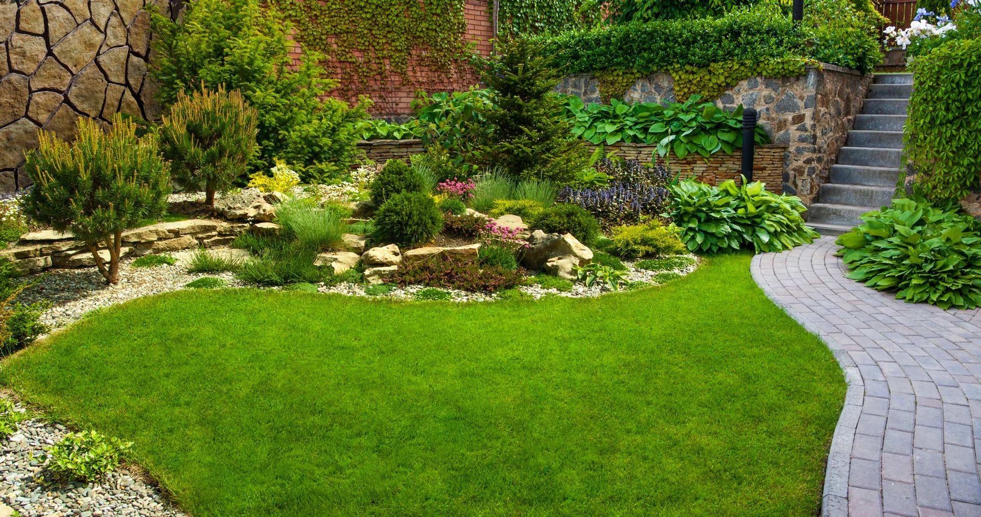 A lush green garden with a brick walkway and stairs leading up to it.