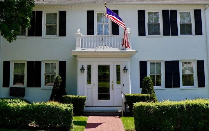 White two-story house with black shutters, an American flag, and a brick walkway, framed by trees and hedges