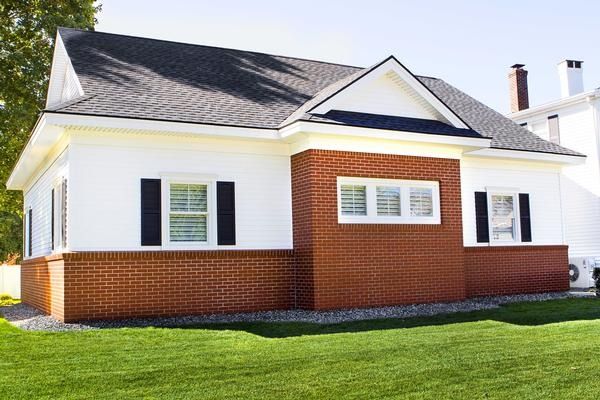 Single-story white house with red brick facade, dark gray roof, and green lawn in sunlight
