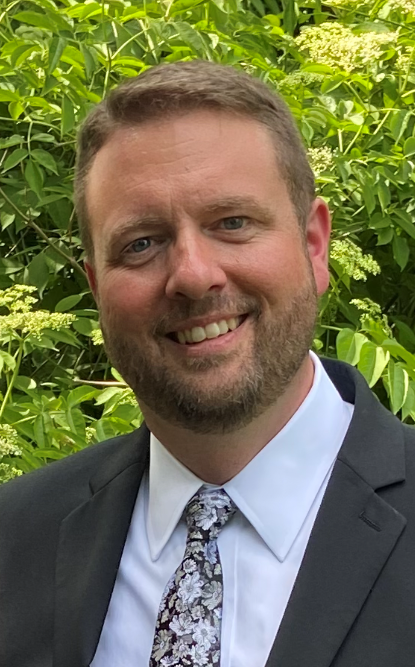 Smiling man in a dark suit and floral tie outdoors in front of green foliage