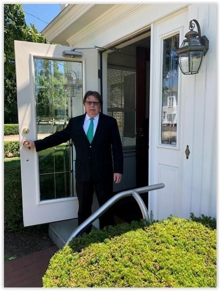 Man in a suit stands in a doorway with the door open, beside a white building and wall lantern.