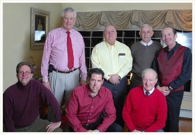 Six people pose indoors in front of a fireplace, wearing colorful dress shirts and sweaters.