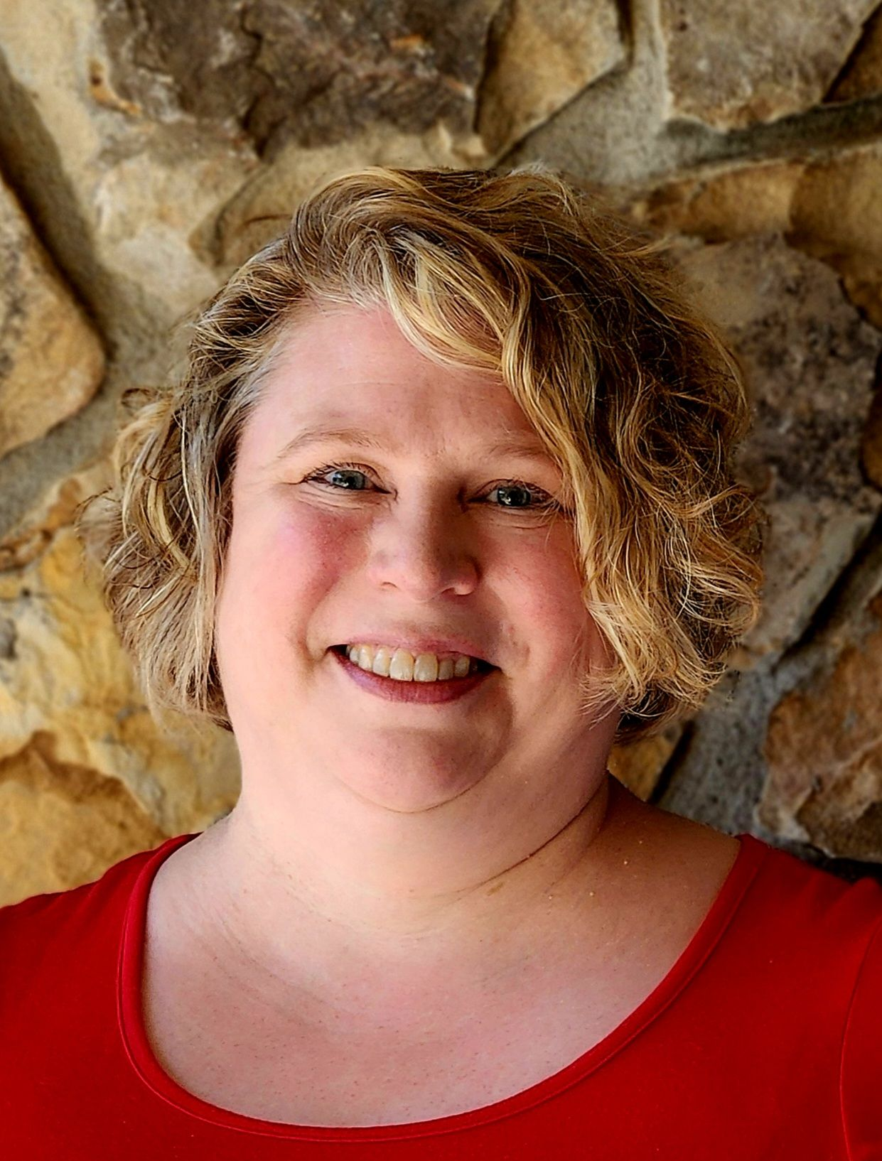 Woman with blonde curly hair smiles at the camera, wearing a green floral top, stone wall background.