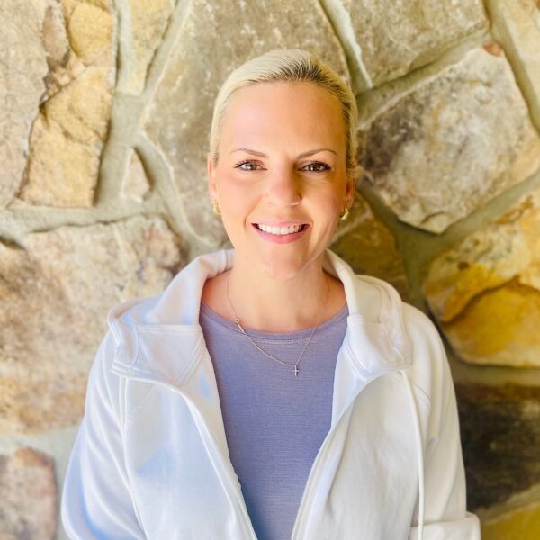 Woman with blonde hair smiles in front of a stone wall, wearing a white jacket and a purple shirt.